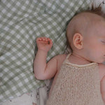 A sleeping newborn baby wearing a cream-colored knit halter dress, lying on a green and white gingham blanket.