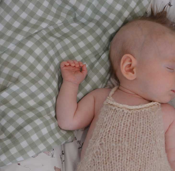 A sleeping newborn baby wearing a cream-colored knit halter dress, lying on a green and white gingham blanket.