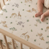 A close-up of a baby's bare legs resting on a cream-colored muslin crib sheet printed with safari animals like giraffes, elephants, and lions, inside a light wood crib.