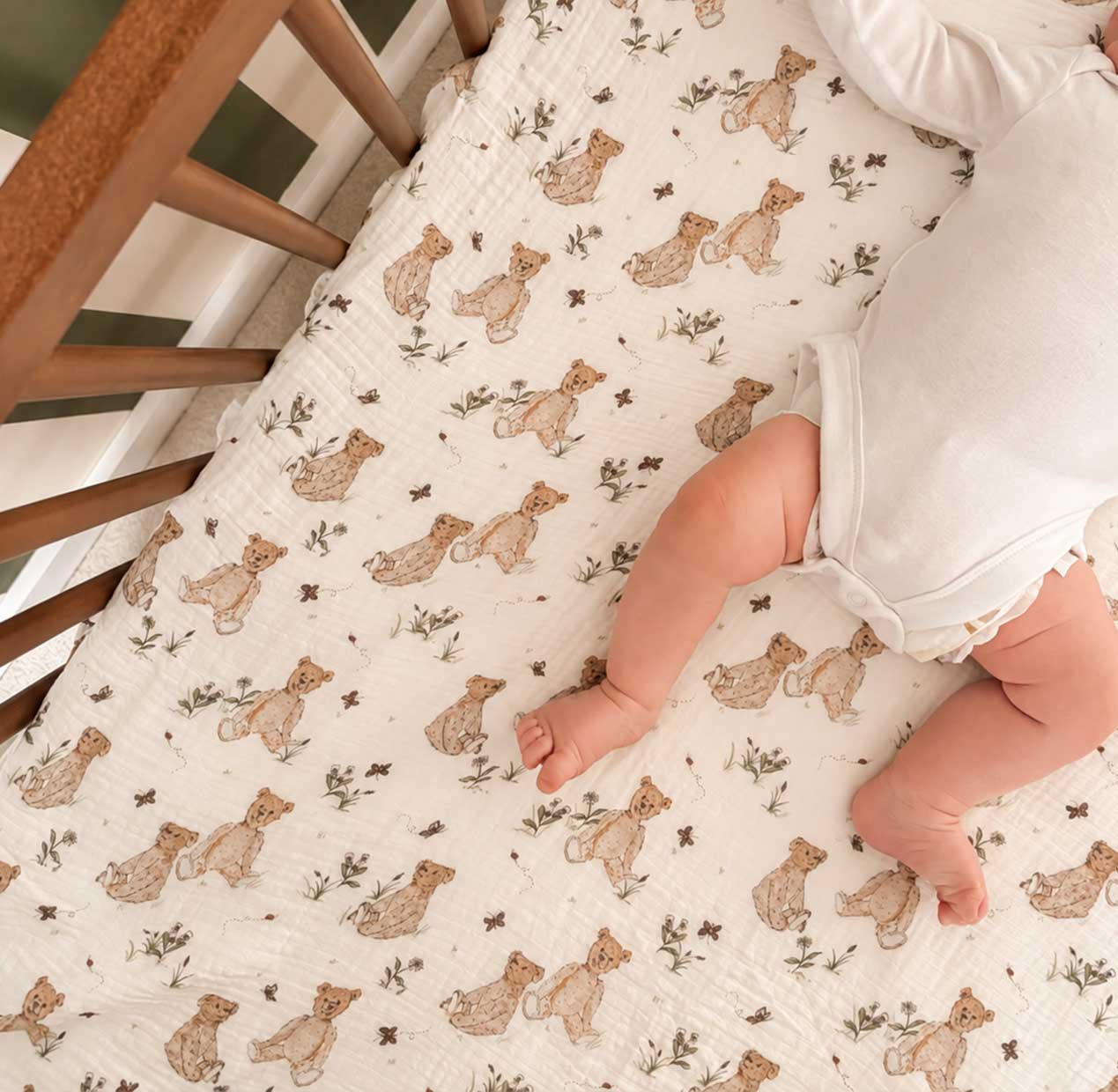 A baby lying on a cream-colored muslin mattress cover printed with a pattern of brown teddy bears and small green leaves, inside a wooden crib.