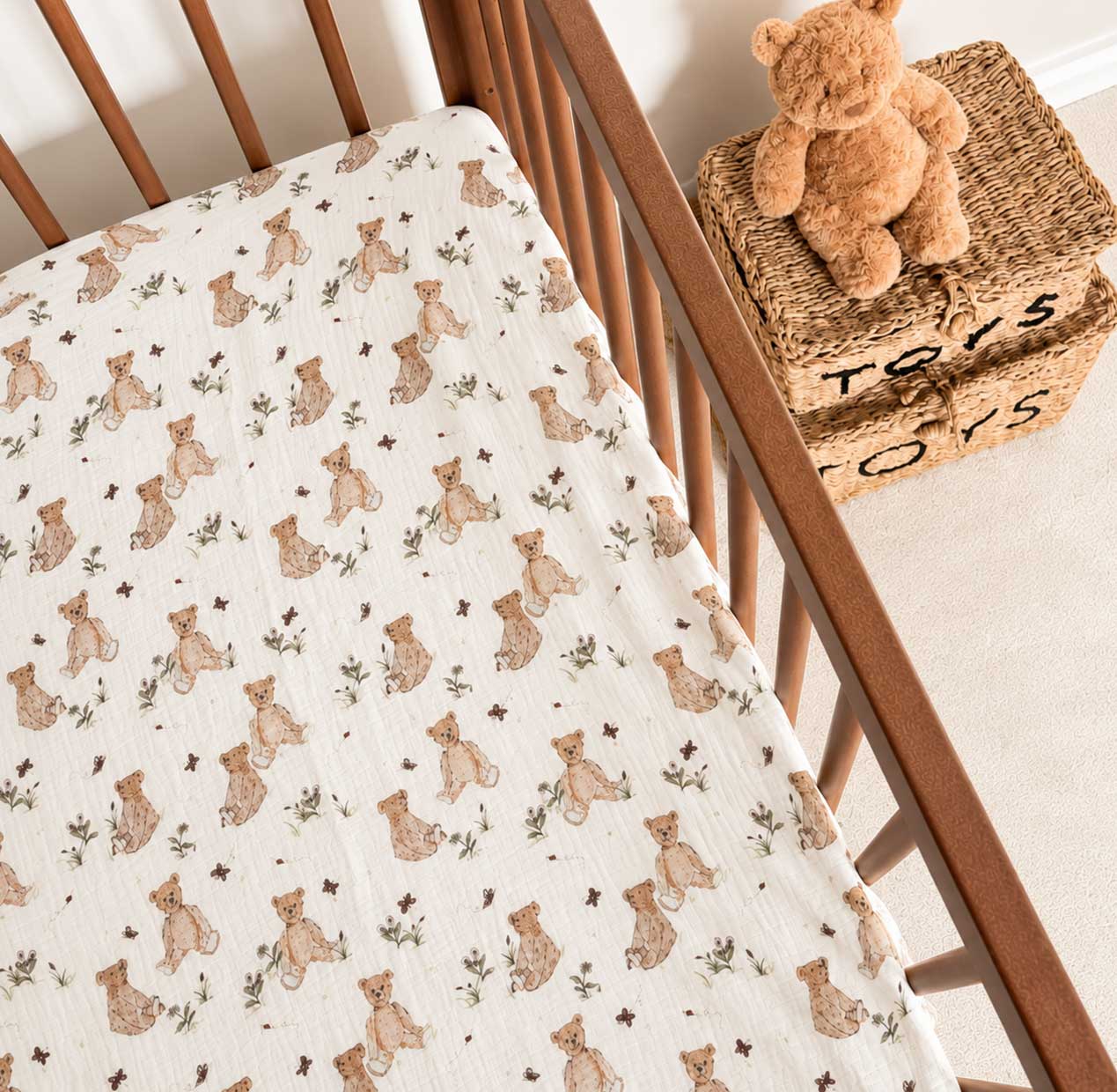 A close-up of a baby blanket or muslin sheet with a repeating print of teddy bears and small stars, laid inside a wooden crib. A woven basket and plush teddy bear sit beside the crib.
