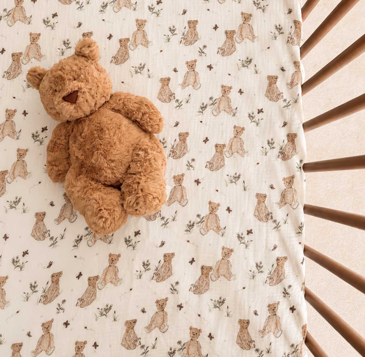 A plush teddy bear rests on a cream-colored crib bedding set printed with small teddy bear motifs, with wooden crib rails visible on the right side.