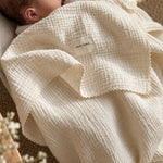 A close-up of a baby wrapped in a textured cream-colored muslin blanket with a visible 'MAMA SHACK' brand label. The baby's head and ear are visible on the left, resting against a soft neutral background.
